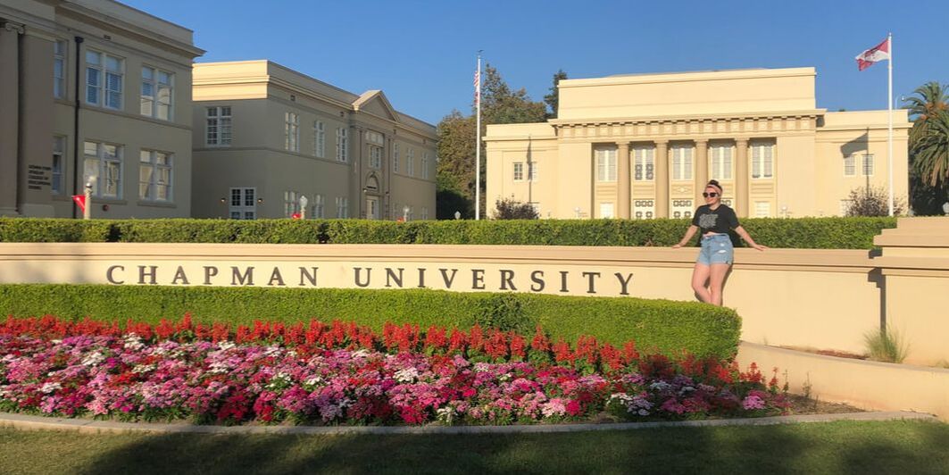 Photo of Ellery Hall in August 2020. She stands in a lush courtyard next to a sign that reads, 